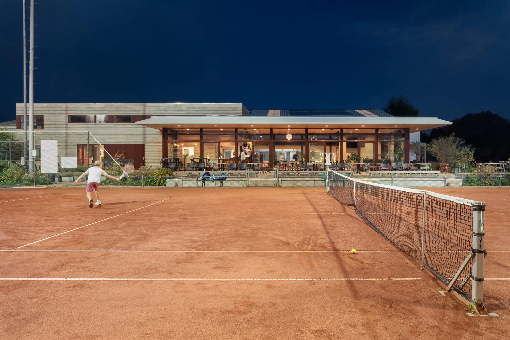 Het verlichte clubhuis in de avond, gezien over de tennisbaan